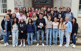 A large group of students and adults participating in the Foley Sister Cities exchange program poses on the stone steps outside a historic building in Hennef, Germany. The group stands in several rows in front of a wooden doorway, with a Hennef sign visible on the building.