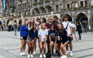 A group of 11 students participating in the Foley Sister Cities exchange program poses in Marienplatz in Munich, Germany, with the ornate Gothic Revival architecture and towering spires of the New Town Hall in the background.