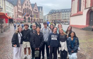 A group of students participating in the Foley Sister Cities exchange program poses for a photo in a historic cobblestone square in Germany, surrounded by traditional European architecture, outdoor cafés and pedestrians.
