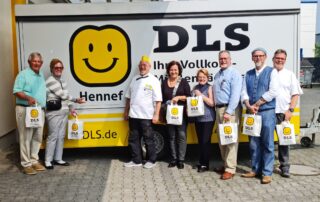 Representatives from Foley and Hennef, Germany, pose for a group photo in front of a DLS Hennef food service trailer featuring a large yellow smiley-face logo. Several participants hold matching DLS gift bags during the visit.